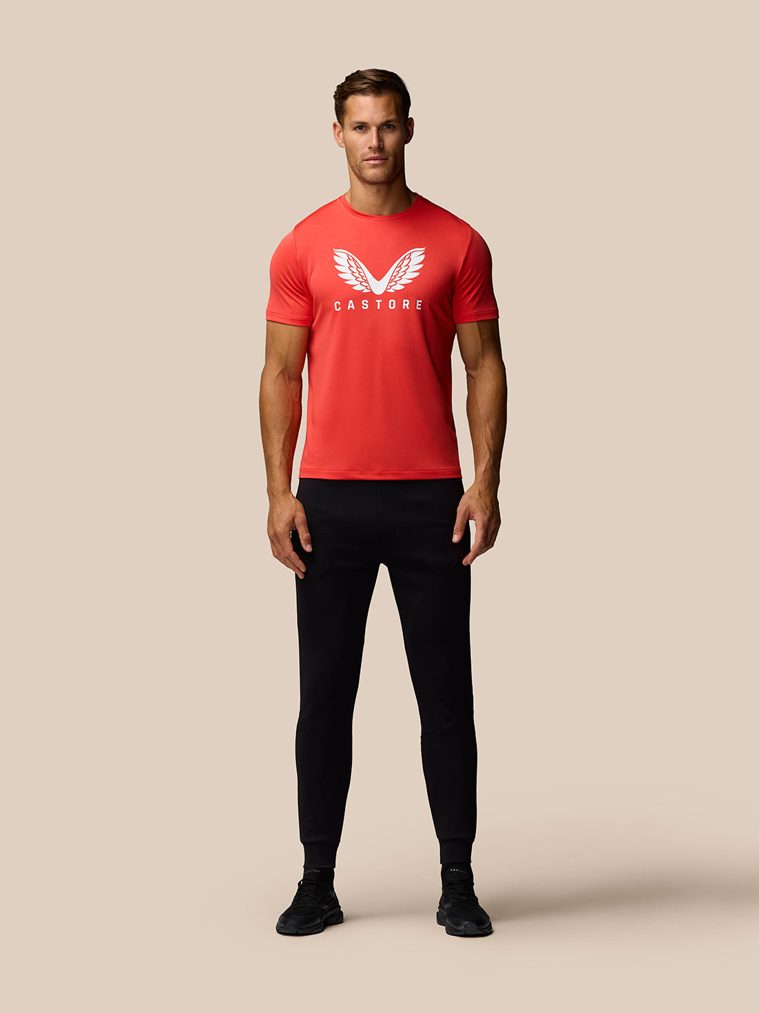 Full body shot of a male model in a poppy red graphic training T shirt with white wing logo and black pants