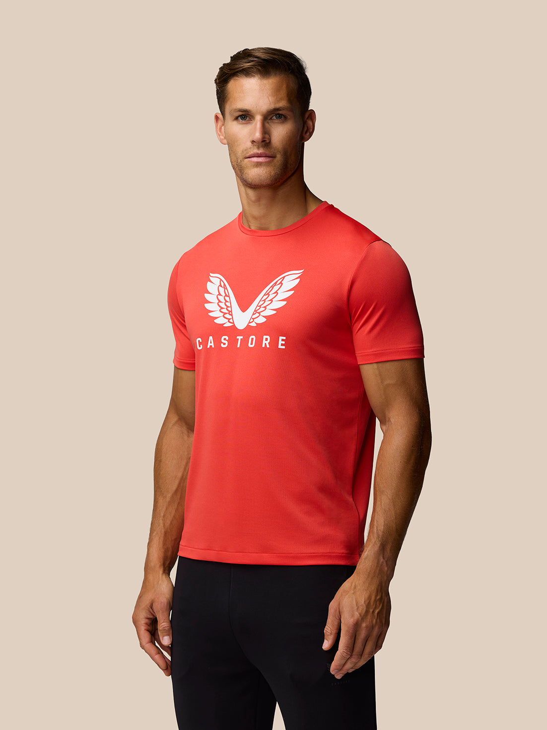 Three quarter view of a male model in a poppy red graphic training T shirt with white wing logo and CASTORE branding