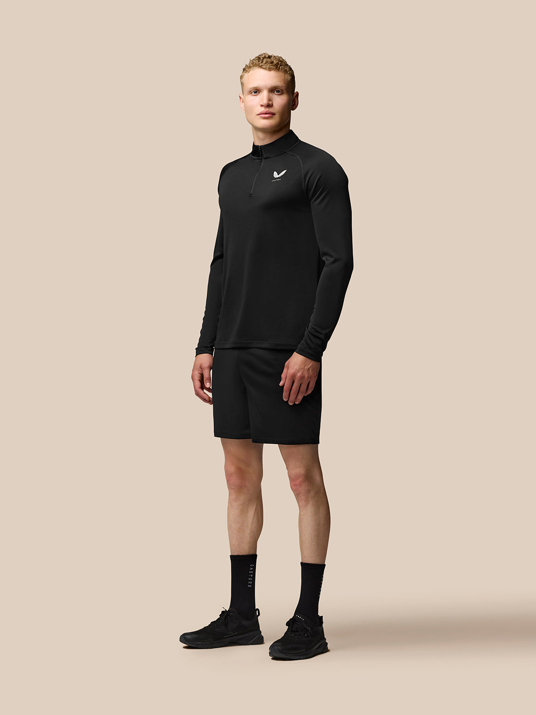 Full body shot of male model in black Adapt Training 1/4 Zip Top with stand collar and logo on chest, paired with shorts