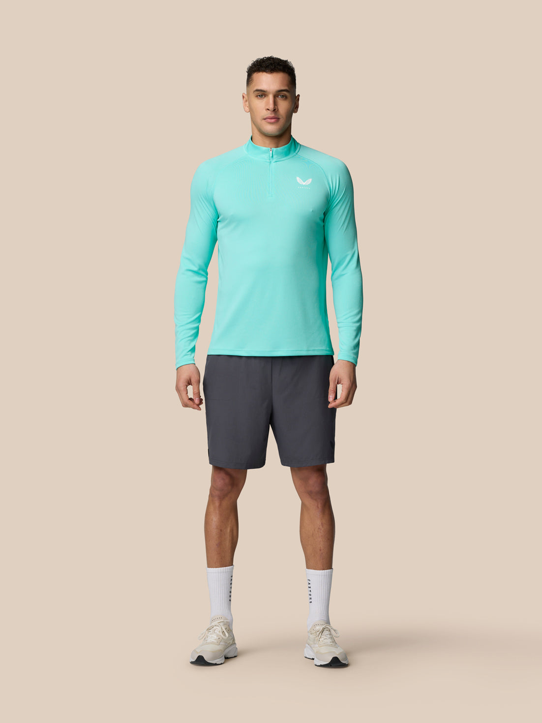 Full-body shot of a man wearing Light Aqua Adapt Training 1/4 Zip Top with dark shorts against a beige background