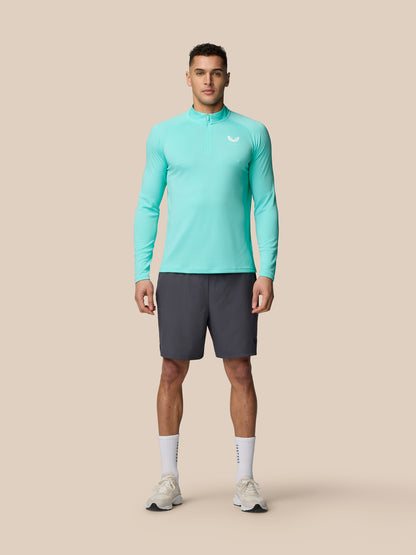 Full-body shot of a man wearing Light Aqua Adapt Training 1/4 Zip Top with dark shorts against a beige background