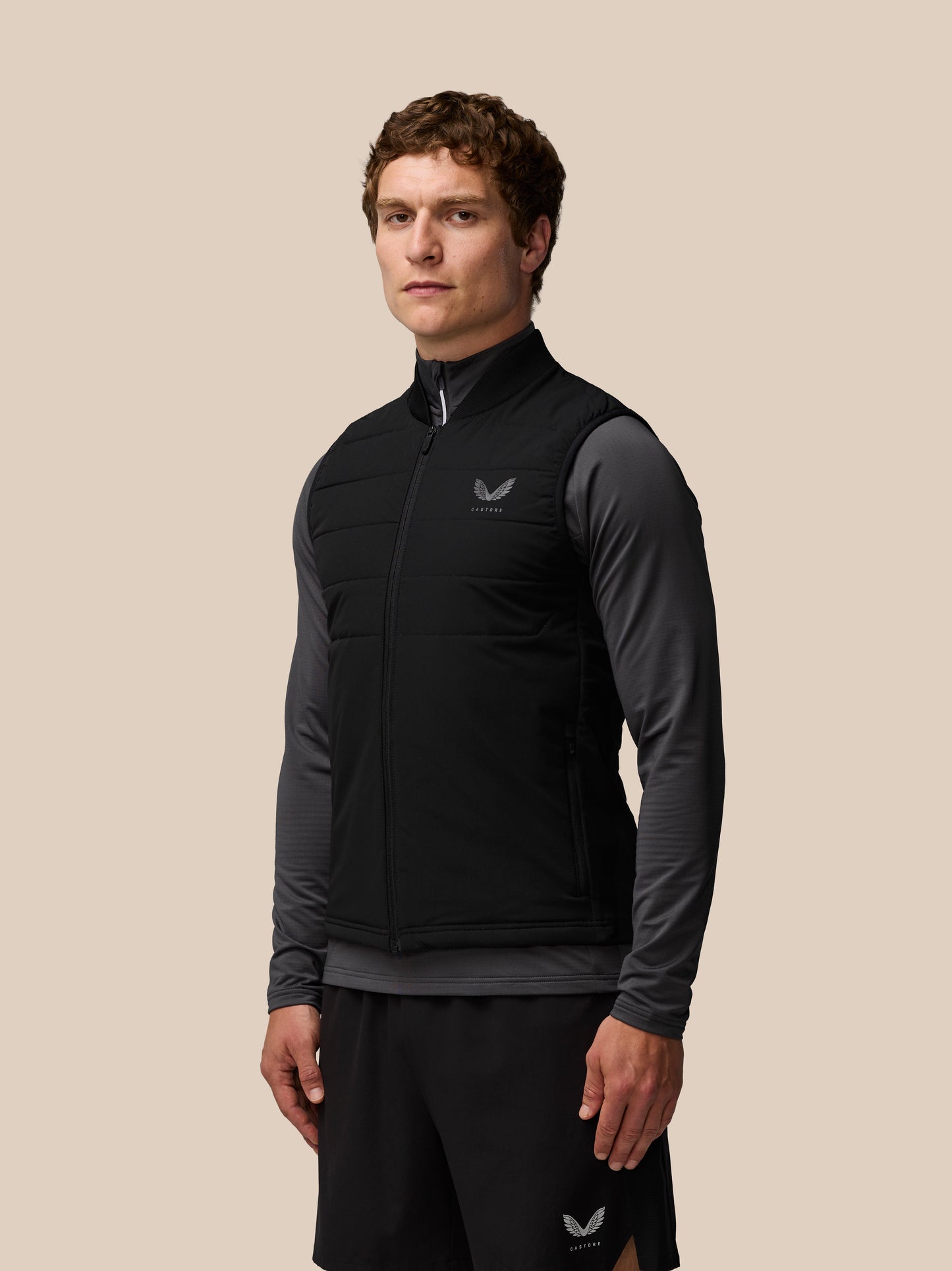 Front view of a man wearing Apex Training Gilet in black, a sleeveless padded vest with zip pockets and a Castore logo on the chest.
