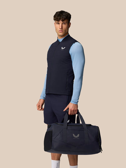 Male model wearing navy Apex Training Gilet over light blue top, carrying a navy Castore gym bag.
