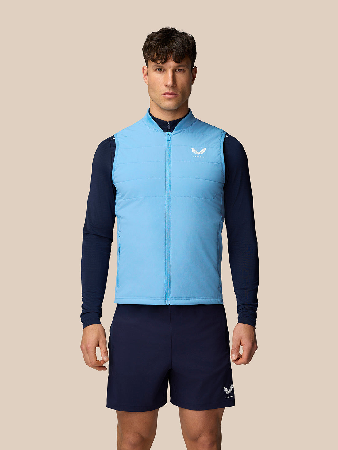 Male model wearing sky blue Apex Training Gilet over a navy base layer and shorts against a beige backdrop.