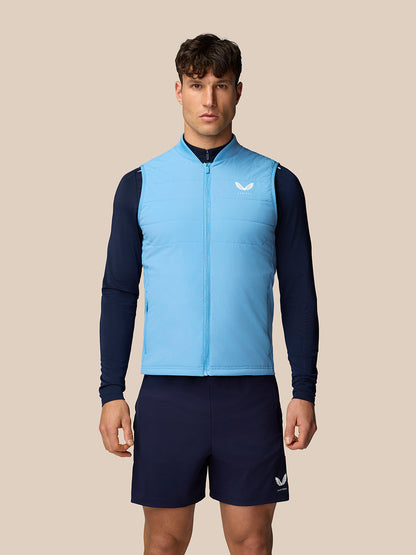 Male model wearing sky blue Apex Training Gilet over a navy base layer and shorts against a beige backdrop.