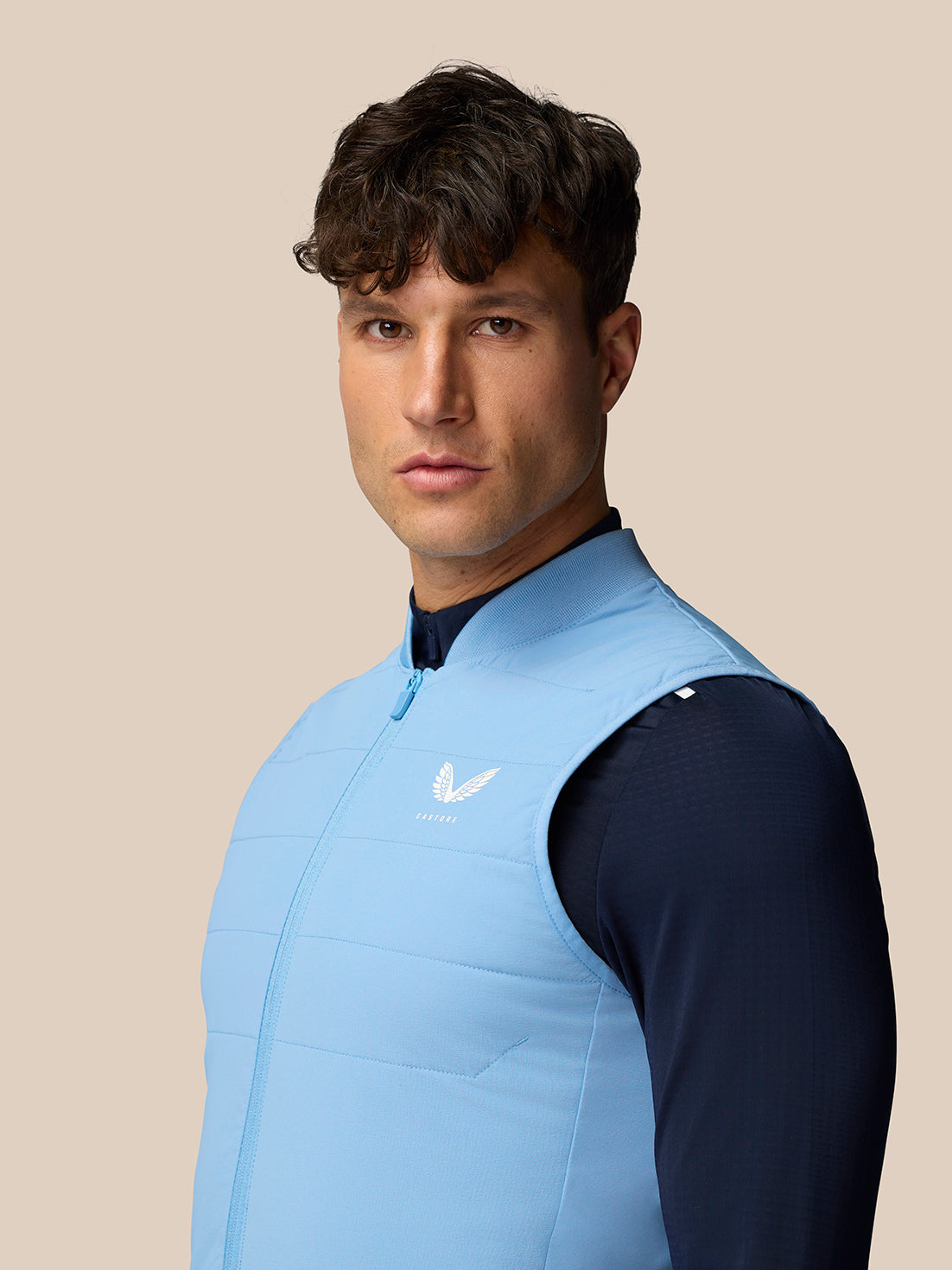 Male model wearing sky blue Apex Training Gilet over navy top, front view with zipper and Castore logo on chest