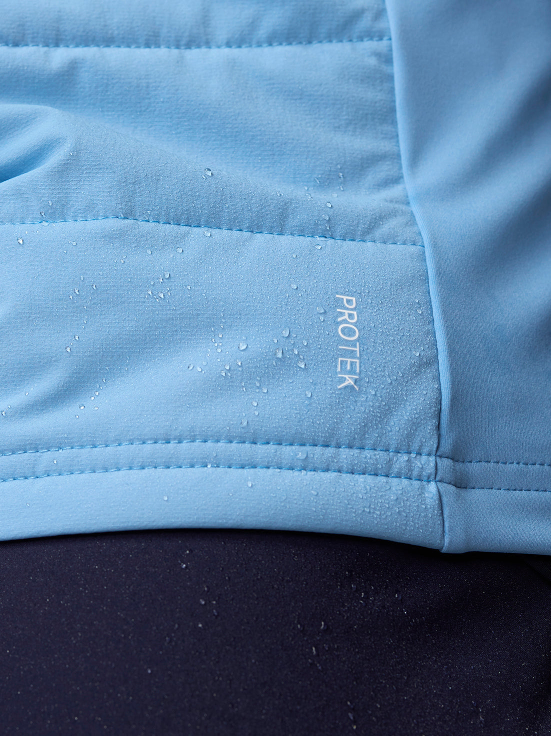 Close-up of sky blue Apex Training Gilet fabric with water droplets and ProTek branding.