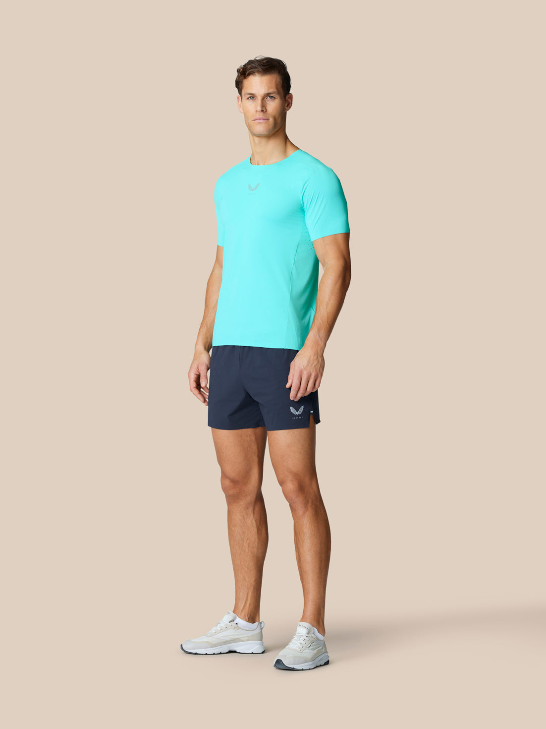 Full body shot of a man in a turquoise top and navy Zone Training Shorts, showcasing the Castore logo on the left thigh