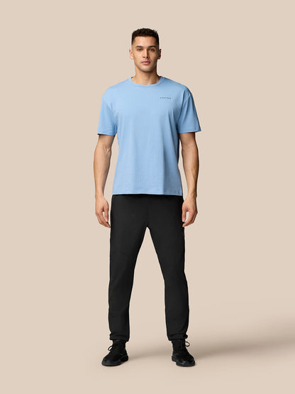 Full-body shot of a man wearing black Flex joggers and a light blue t-shirt against a beige background