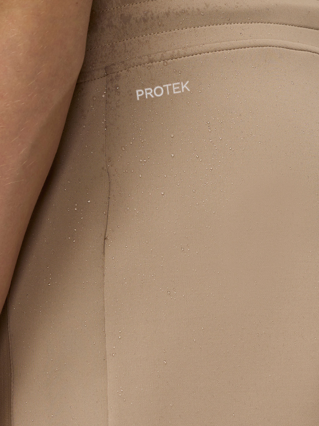 Close up of beige jogger fabric with ProTek logo and water droplets highlighting water resistance