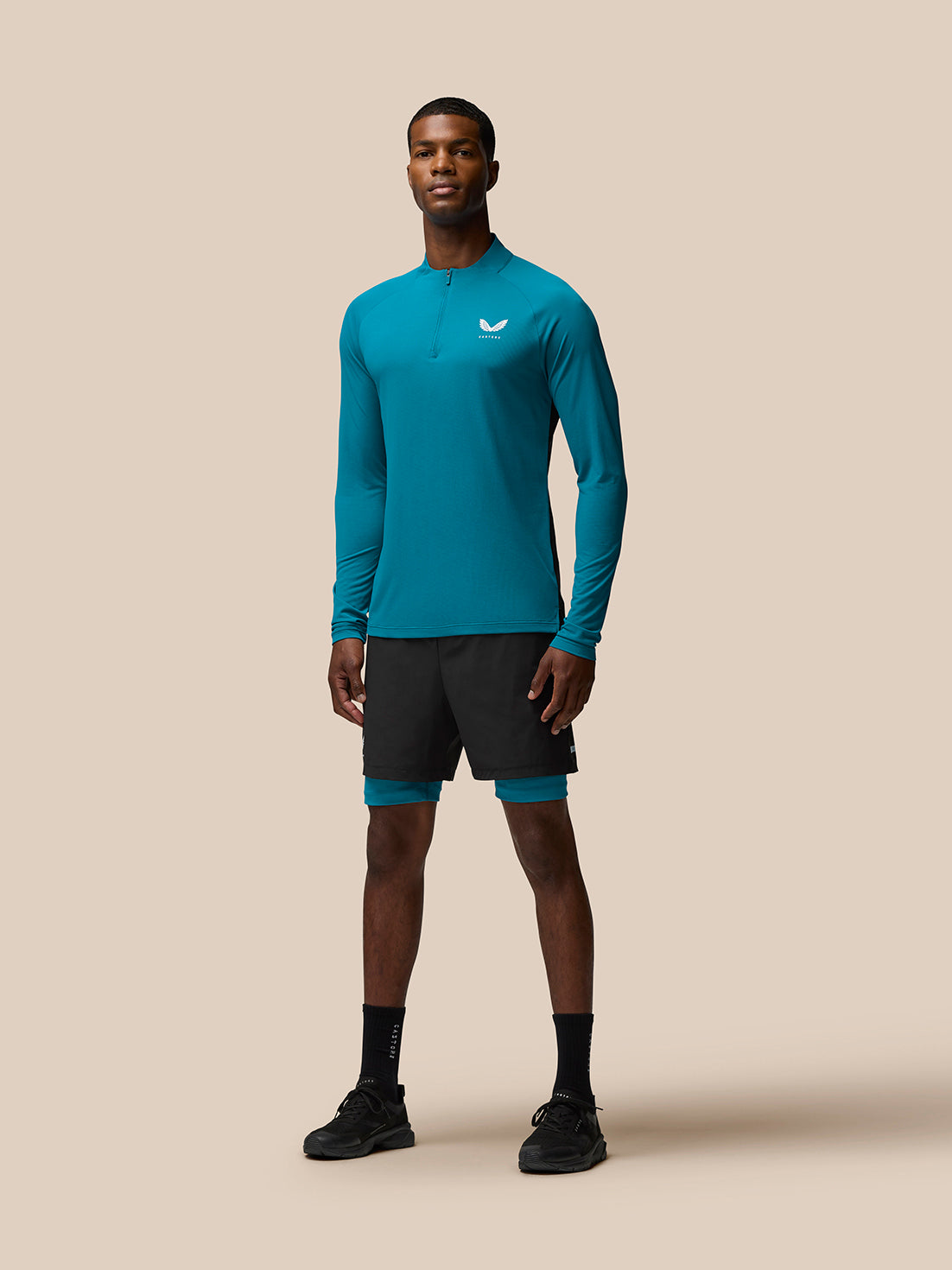 Full-body front view of a man wearing teal Flow Training top with quarter zip and left chest logo against a beige background