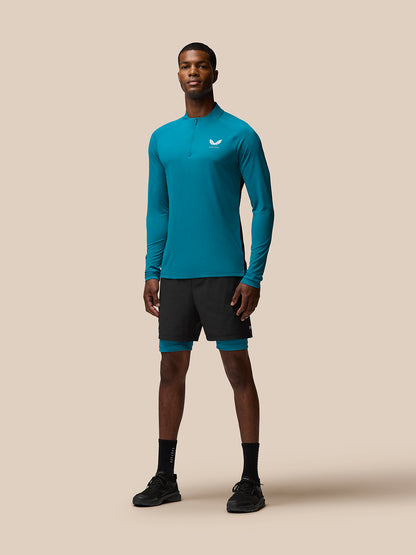 Full-body front view of a man wearing teal Flow Training top with quarter zip and left chest logo against a beige background