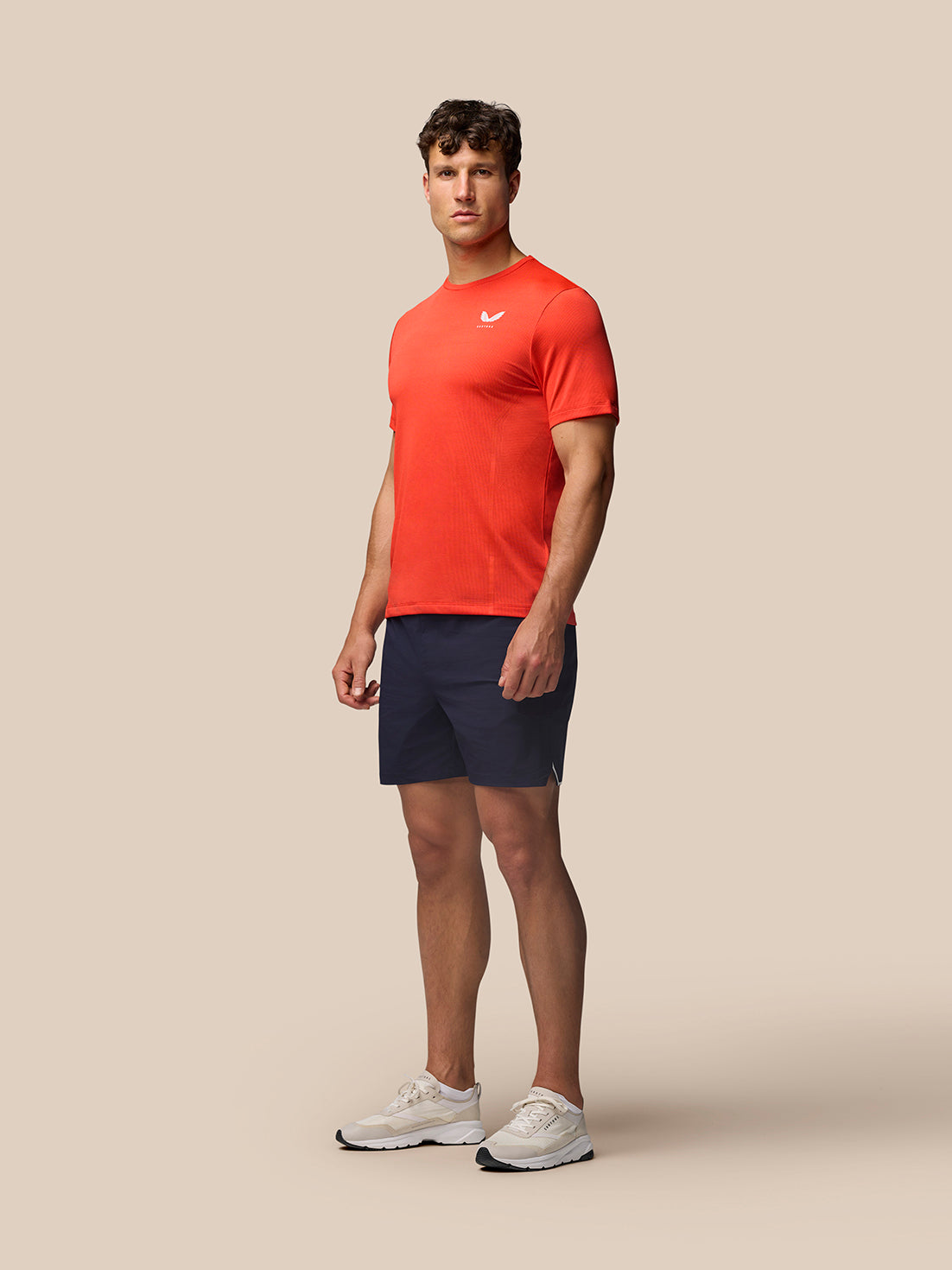 Male model wearing red Apex Training T-Shirt with white Castore branding on the chest, navy shorts and white sneakers.