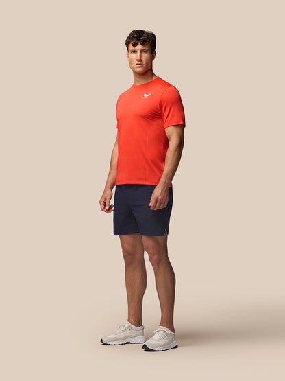 Male model wearing red Apex Training T-Shirt with white Castore branding on the chest, navy shorts and white sneakers.