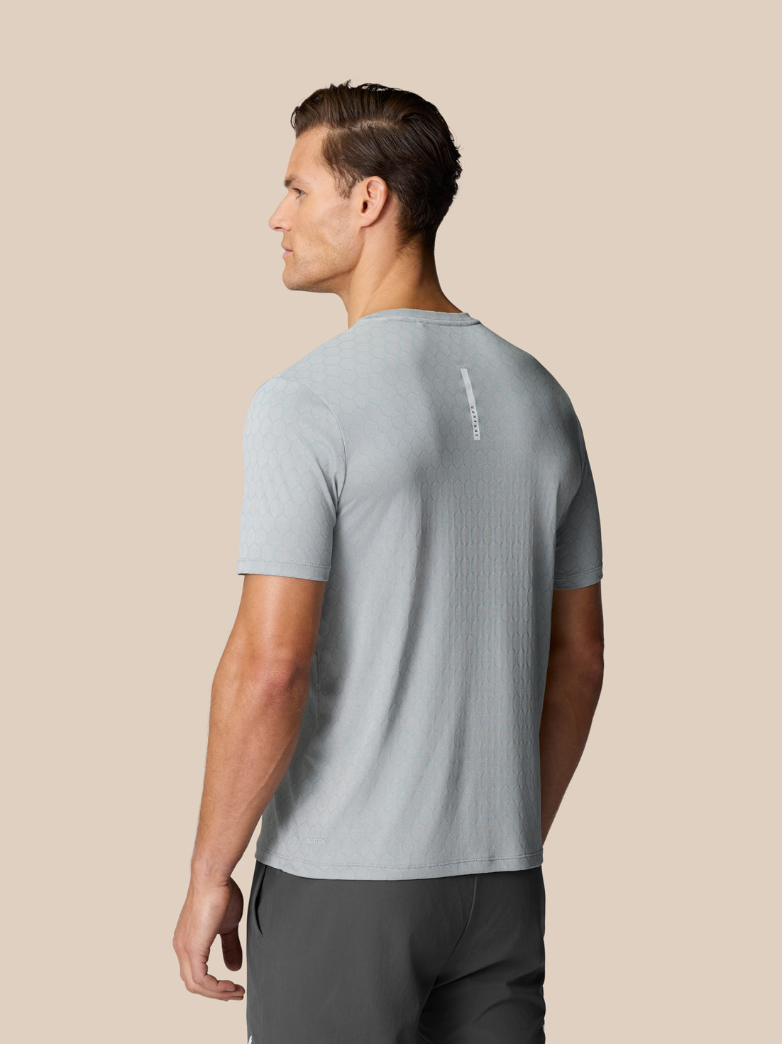 Back view of a man wearing a light grey Flow Training T Shirt with a hexagonal knit pattern and a vertical Castore tag on the upper back.