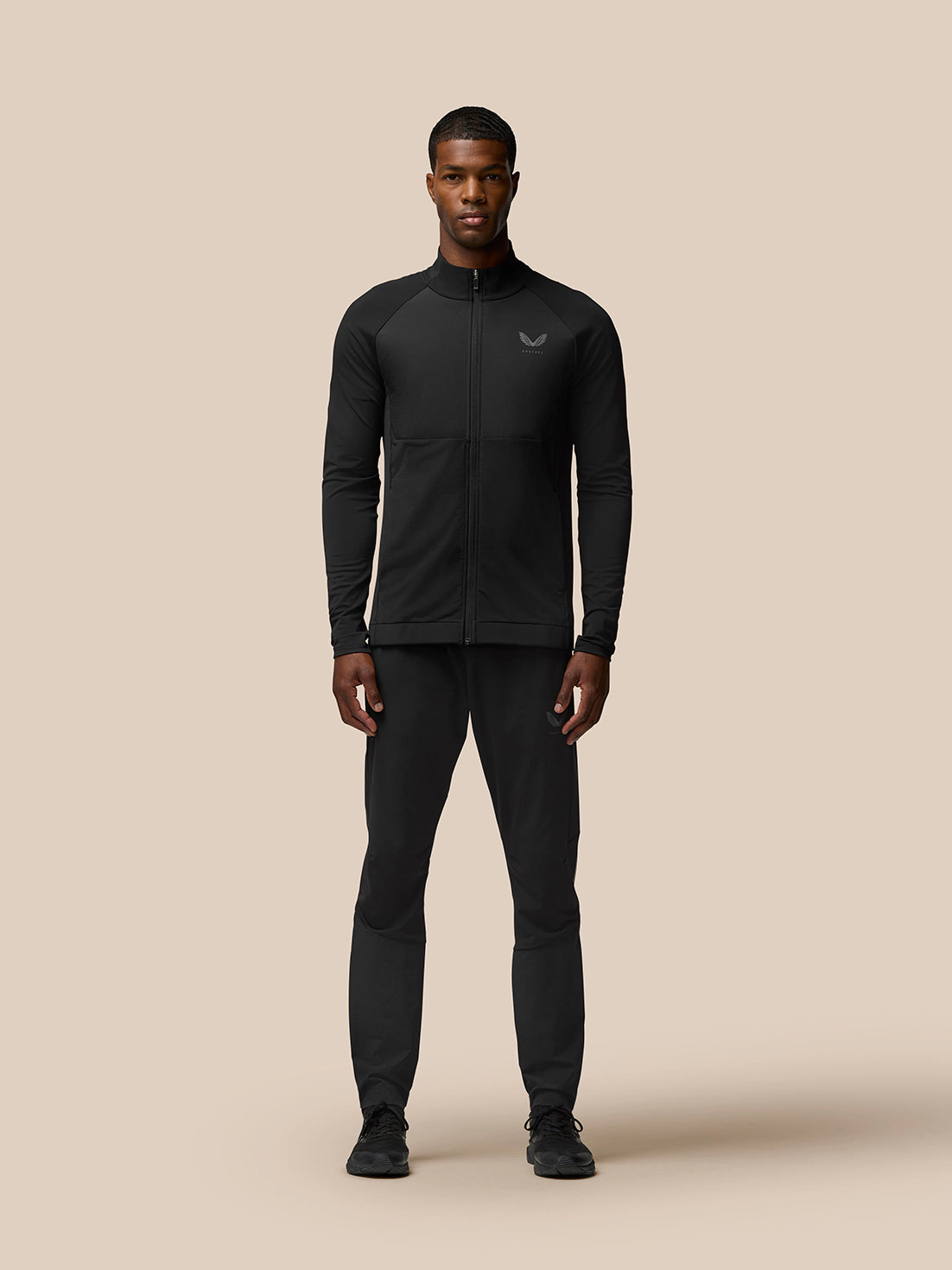 Full body shot of a male model wearing Flow Training Jacket black with matching pants against a beige backdrop