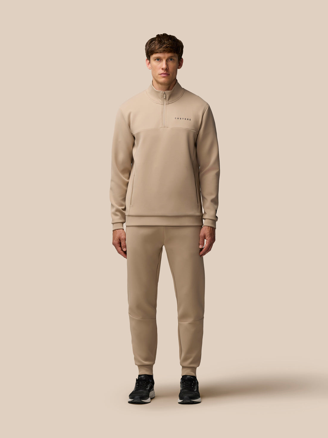 Full-body view of a model in a mushroom beige Flex Lifestyle tracksuit with a center front quarter zip and zipped pockets.