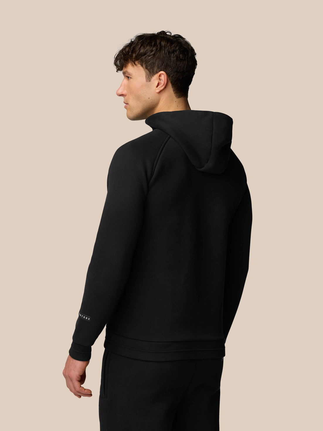 Back view of a man in black Adapt Fleece Zip-Through Hoodie with hood up and sleeve logo