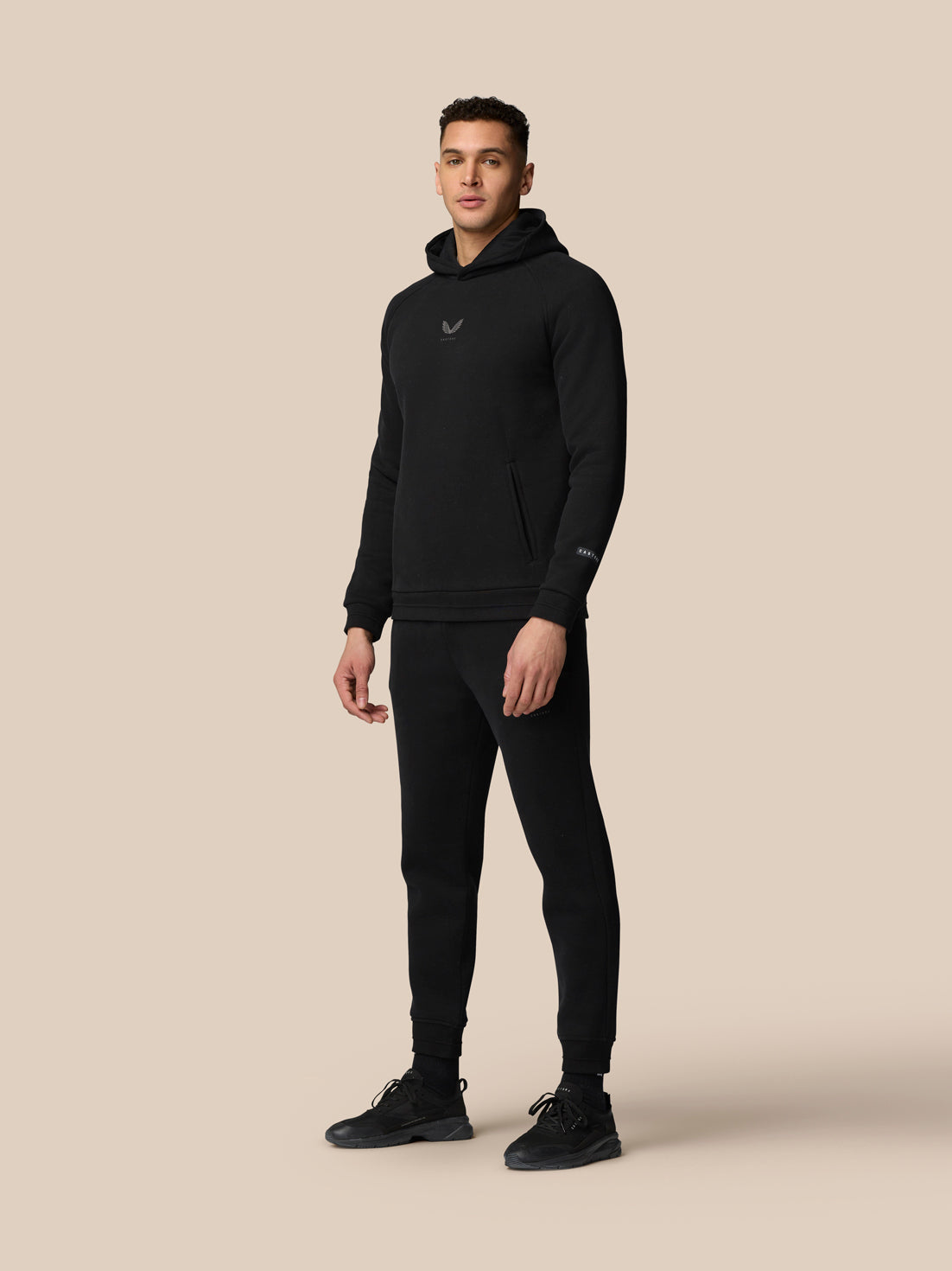 Full-body shot of model in black Adapt Fleece Hoodie with matching joggers and black sneakers against a beige background.