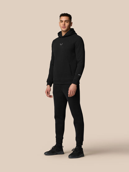 Full-body shot of model in black Adapt Fleece Hoodie with matching joggers and black sneakers against a beige background.