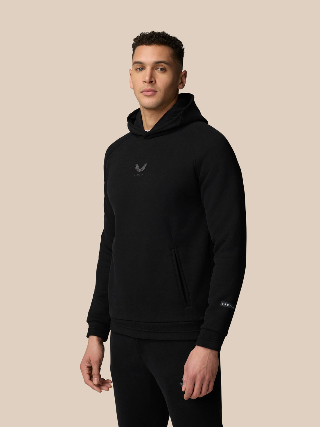 Male model wearing black Adapt Fleece Hoodie with hood and front pocket against a beige background.