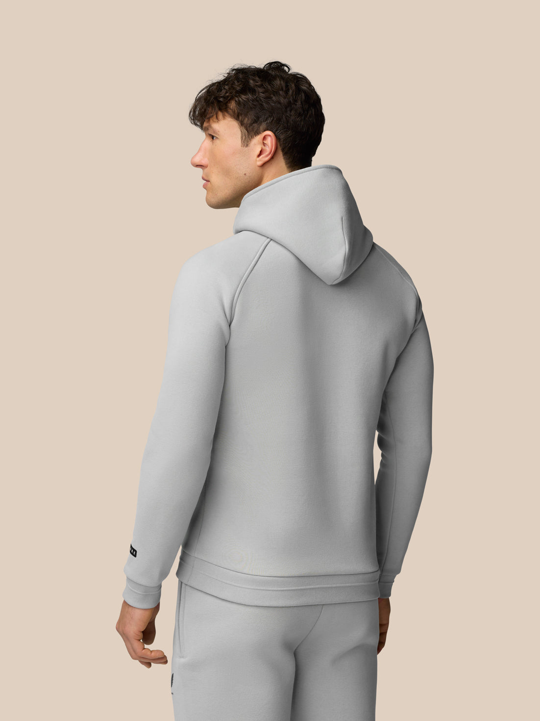 Back view of a man wearing a light grey Adapt Fleece Hoodie with hood up against a beige background.