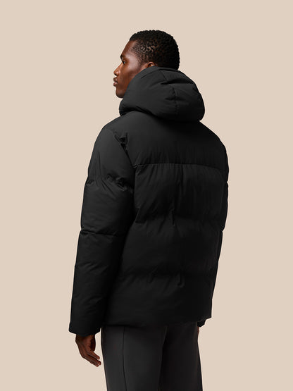 Rear view of a black puffer jacket worn by a model, hood up and quilted sections visible.