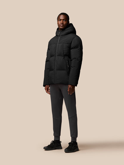 Full-body portrait of a man in a black Castore puffer jacket with hood down, paired with dark joggers and sneakers