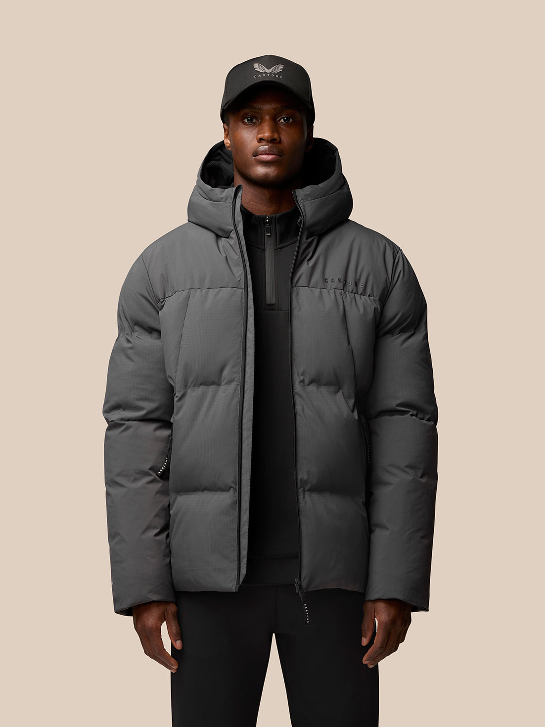 Model wearing charcoal puffer jacket with hood, front zip and zipped pockets; Castore logo on left chest and hood.
