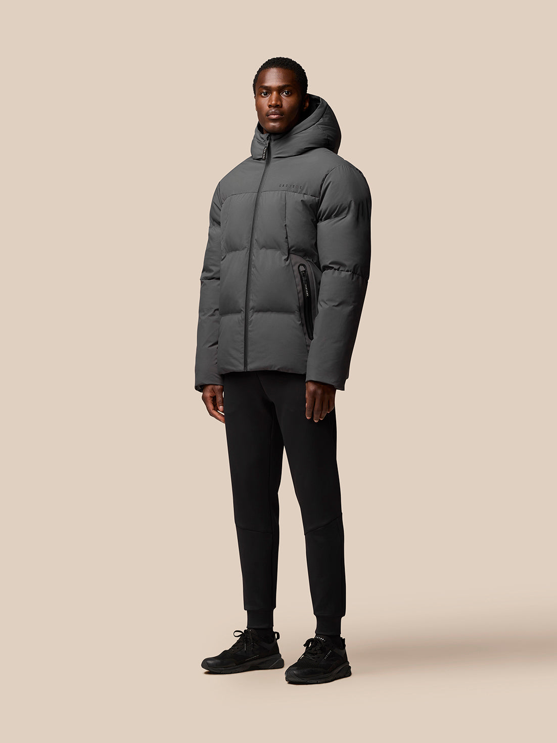 Male model in a charcoal hooded puffer jacket with zip front and side pockets; Castore branding on chest and cap.