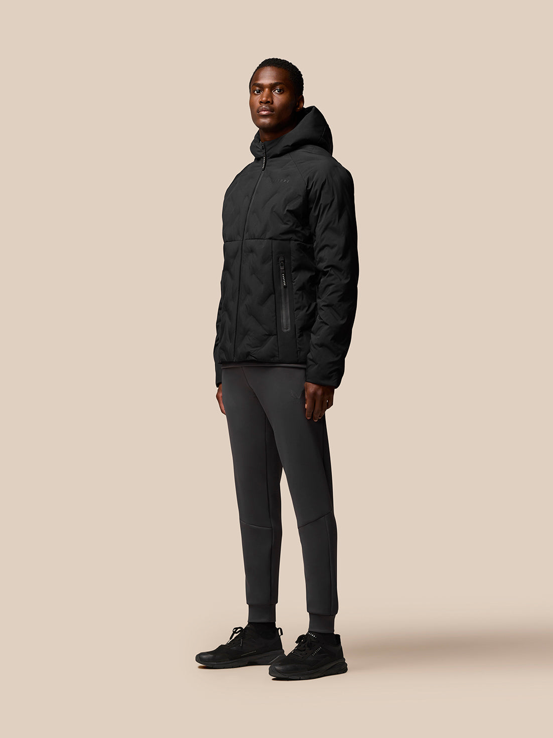 Full-body side view of a man in a black padded Castore jacket with hood paired with dark joggers and sneakers