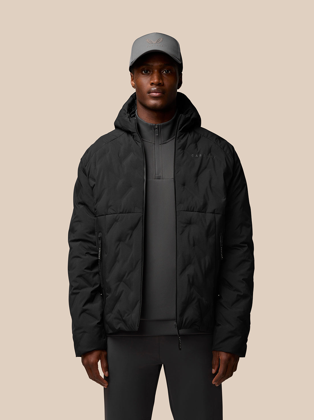 Male model wearing black padded jacket with hood front zip side pockets Castore logo on chest and ProTek branding on sleeve