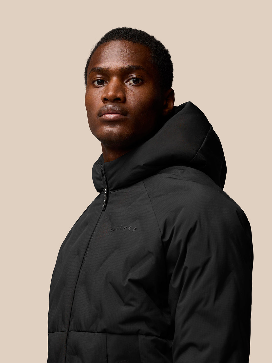 Model wearing black padded hooded jacket with front zip and external zipped pockets; Castore logo on chest and ProTek branding on sleeve