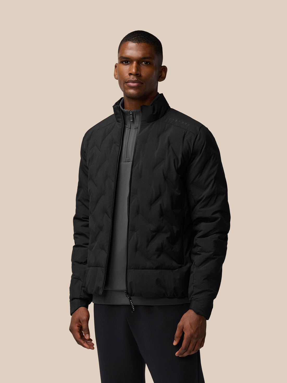 Male model wearing black padded jacket with stand collar and front zip against a beige studio background