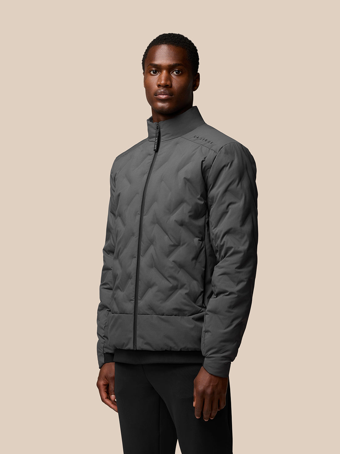 Male model wearing charcoal padded jacket with stand collar and front zip, highlighting Castore and Protek branding.