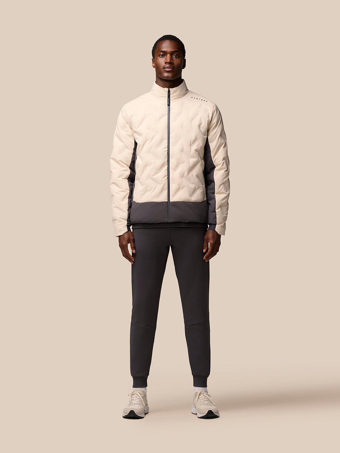 Full-body front view of a man wearing a beige two-tone padded jacket with dark lower panel