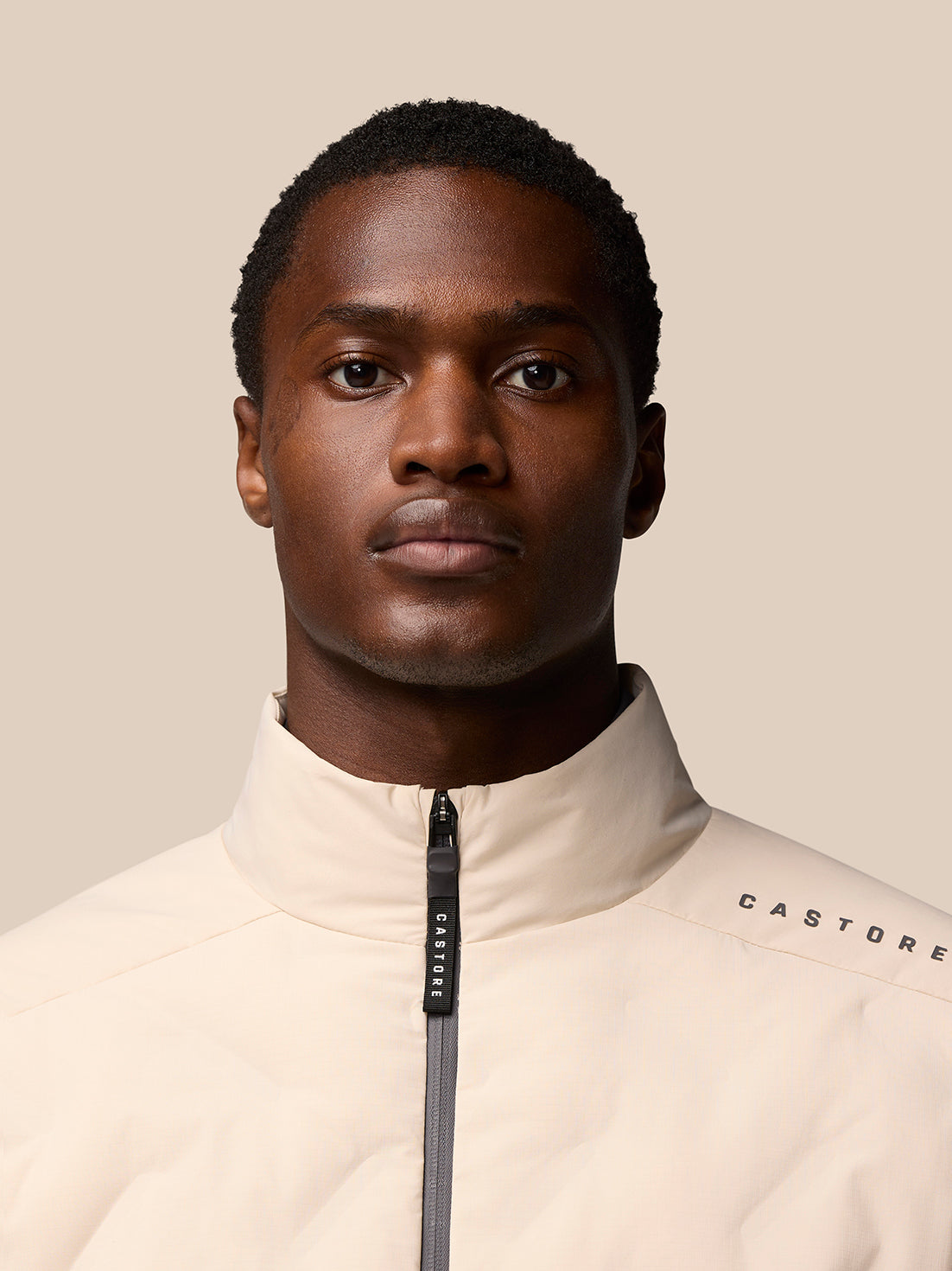 Male model wearing beige padded jacket with center zip and Castore branding on the shoulder