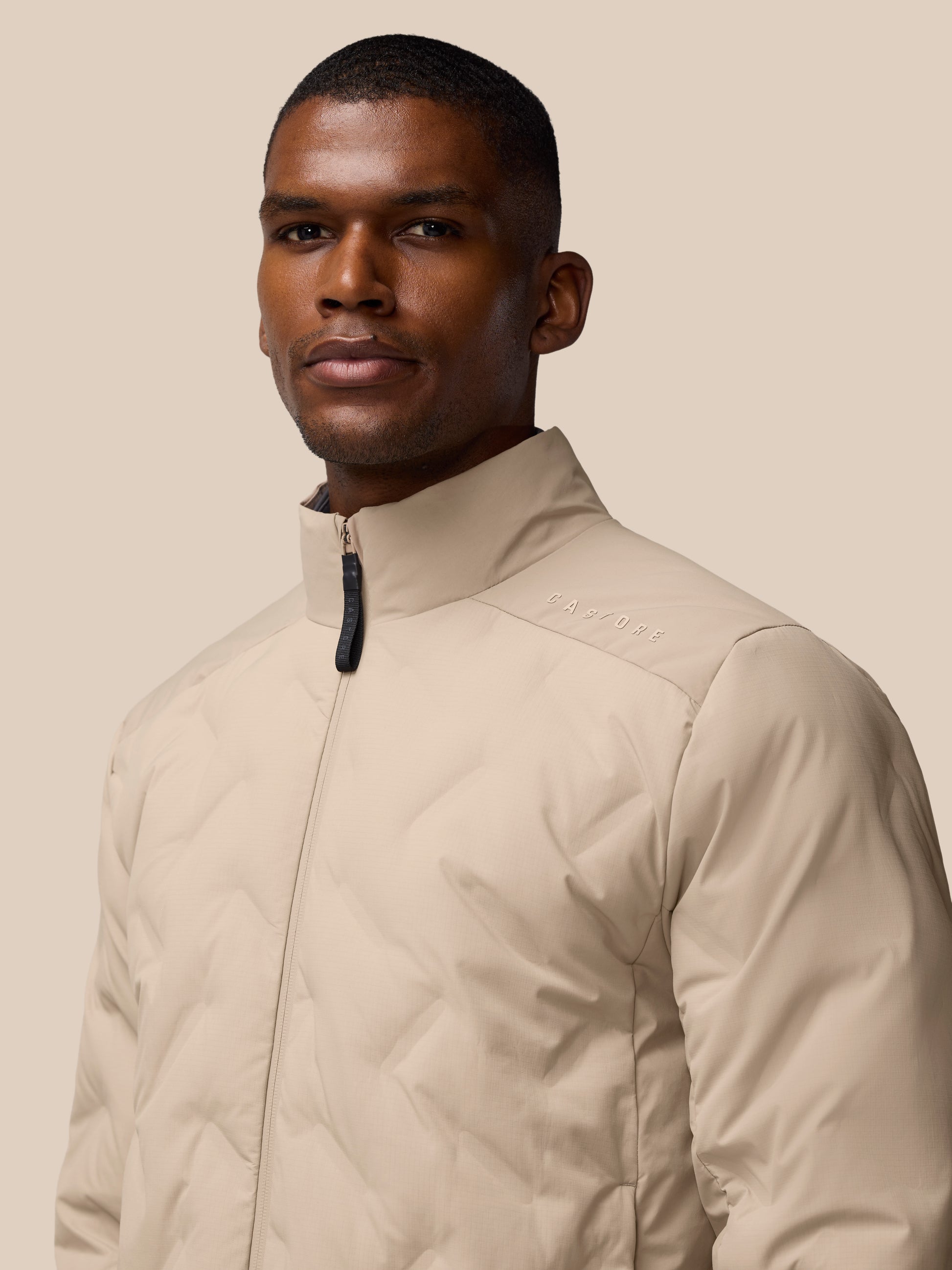 Portrait of a man wearing a mushroom colored padded jacket with a stand-up collar and front zipper.
