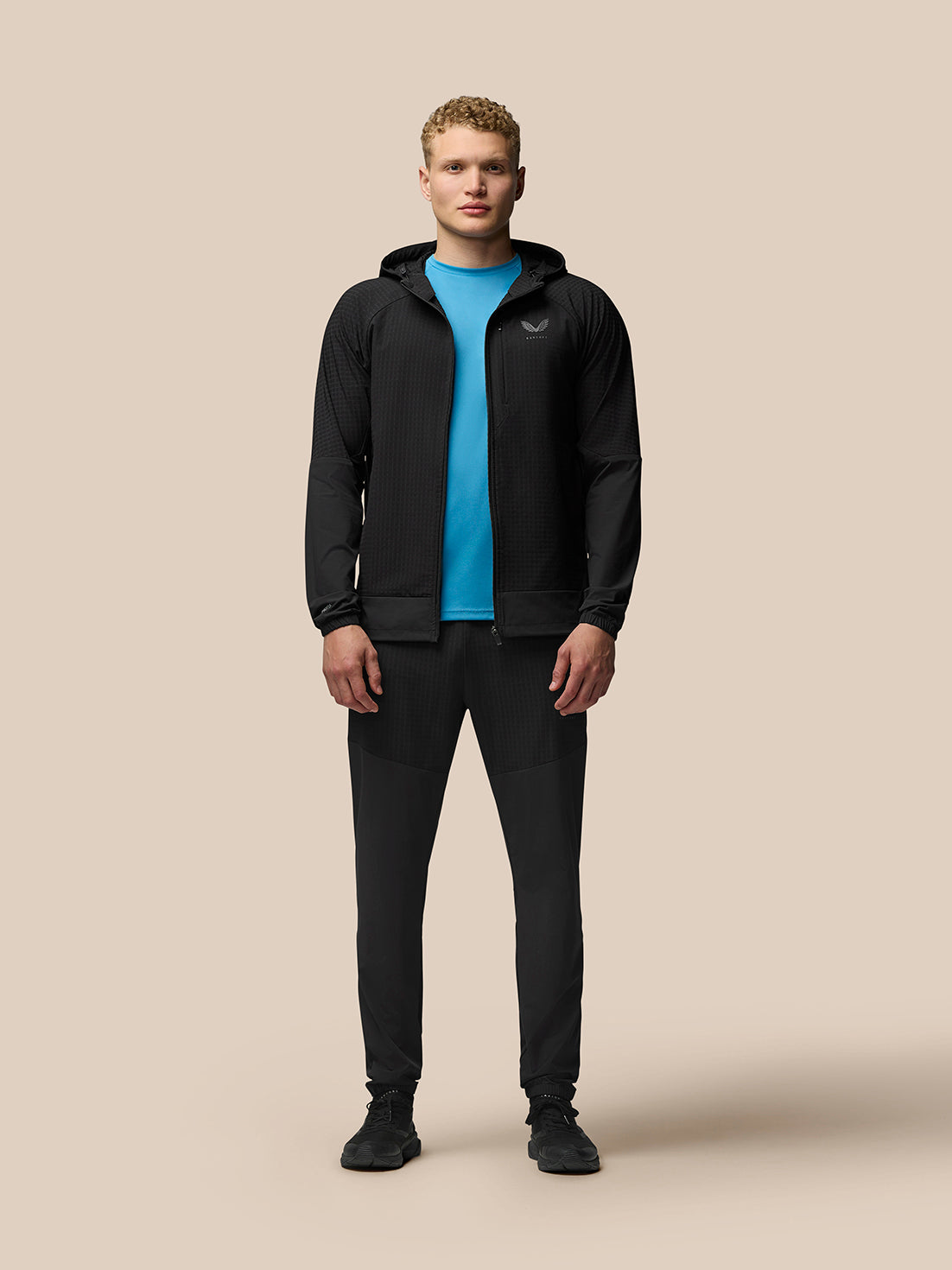 Full body view of a male model in a black hooded Flex Winterised Jacket over a bright blue top and black pants