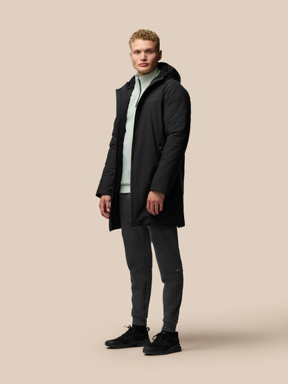 Full body shot of a male model in a black longline hooded jacket zipped up over a pale green top against a beige background
