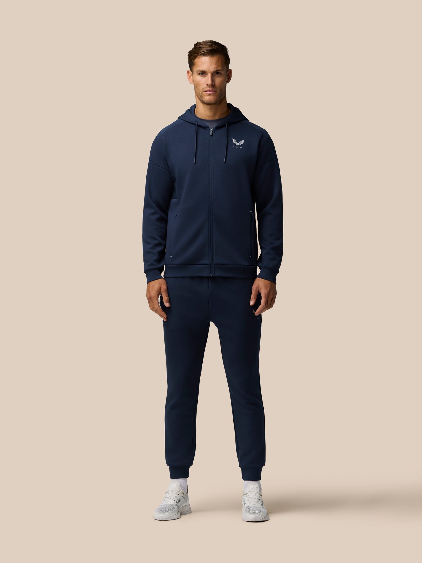Male model wearing navy travel lifestyle jogger set with hooded zip jacket and matching pants against a beige background