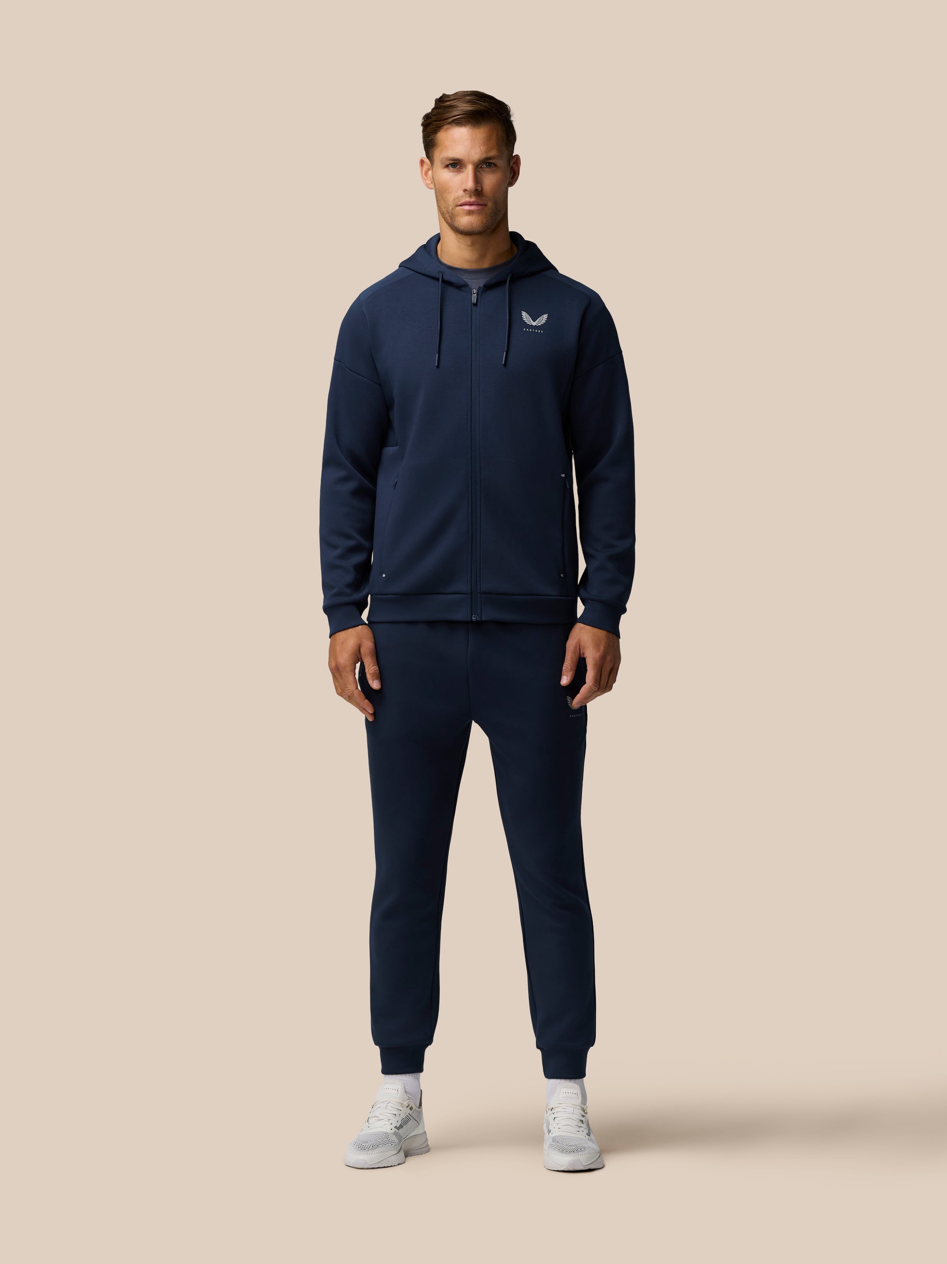 Male model wearing navy travel lifestyle jogger set with hooded zip jacket and matching pants against a beige background