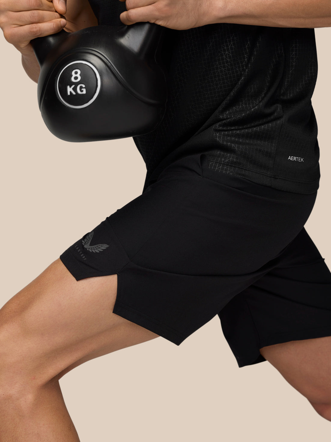 Side view of a person wearing black Adapt Training Shorts with AERTEK branding on the thigh while lifting a kettlebell.