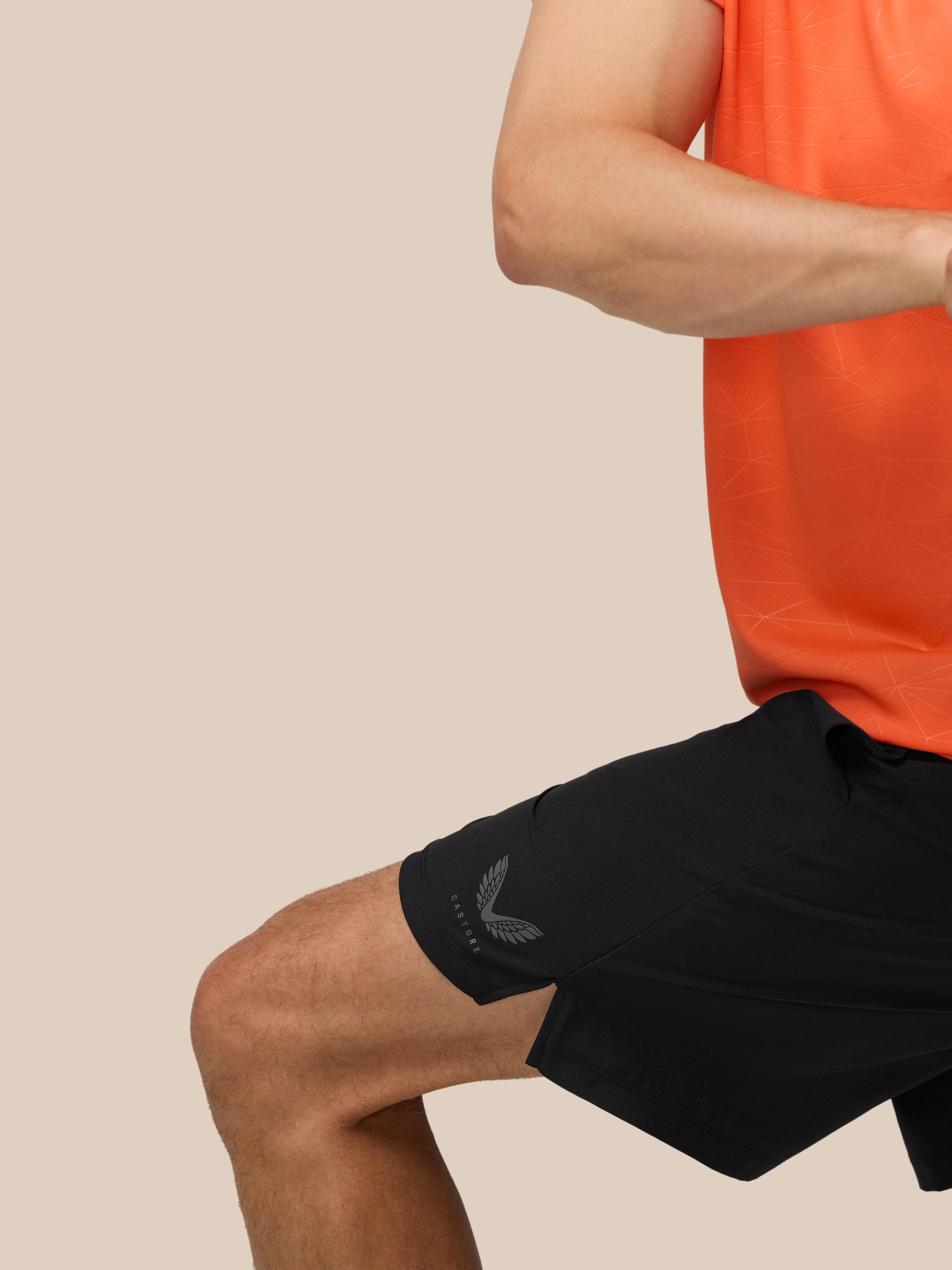Partial view of a person in orange athletic shirt and black shorts with a logo on the left thigh against a beige background.