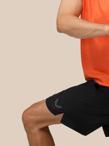 Partial view of a person in orange athletic shirt and black shorts with a logo on the left thigh against a beige background.