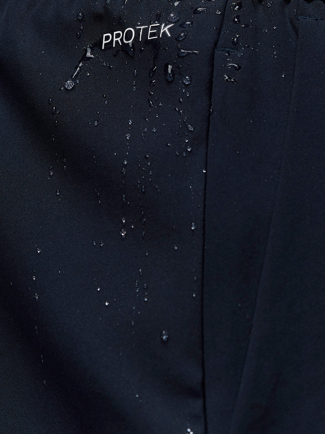 Close-up of midnight navy Adapt Training Short fabric showing water beading and PROTEK branding.