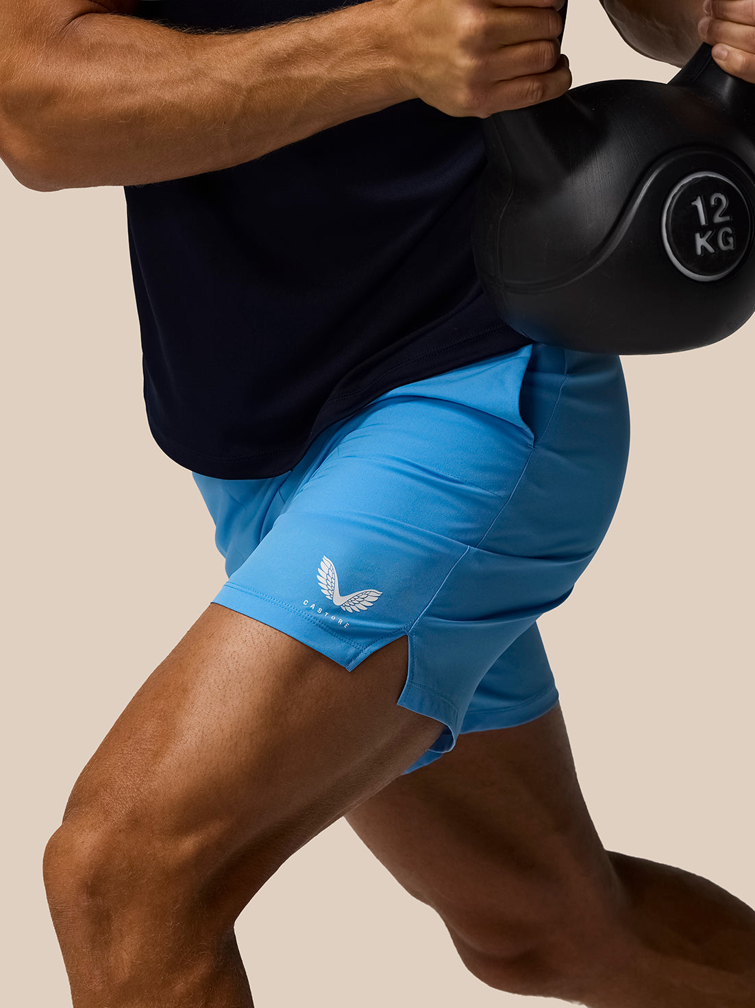 Close-up of a lifter wearing cool blue Adapt Training Shorts with a white Castore wing logo, gripping a 12 kg kettlebell.