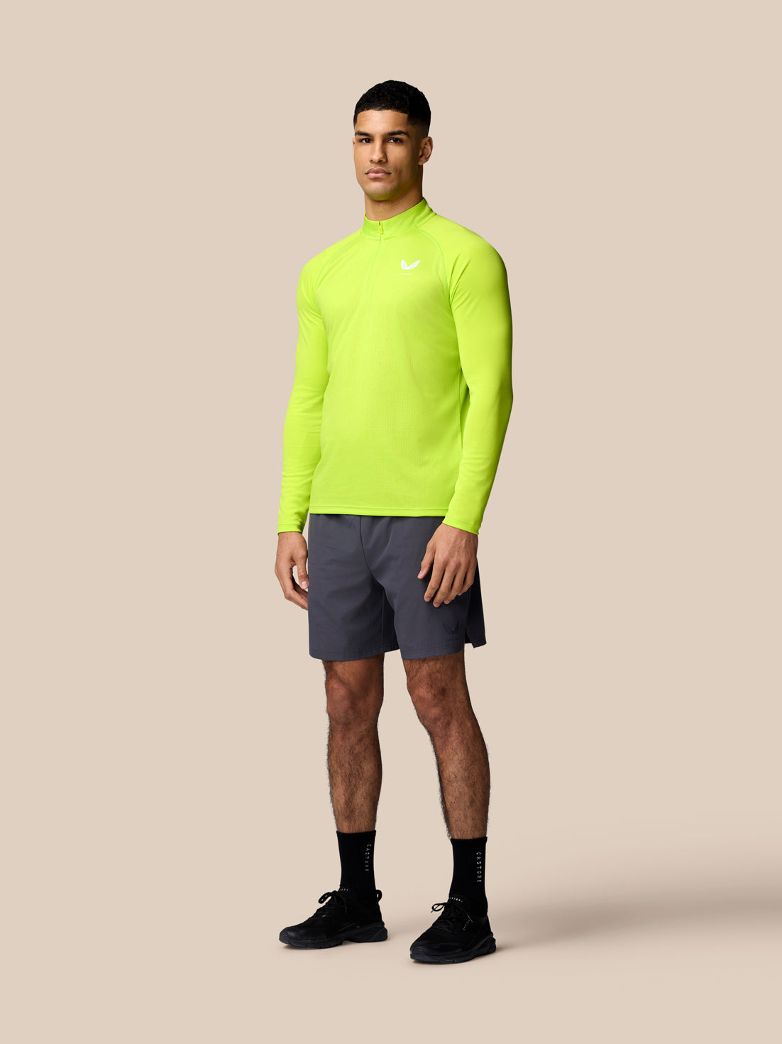 Male model wearing a neon lime long sleeve training top and gunmetal shorts against a beige backdrop.