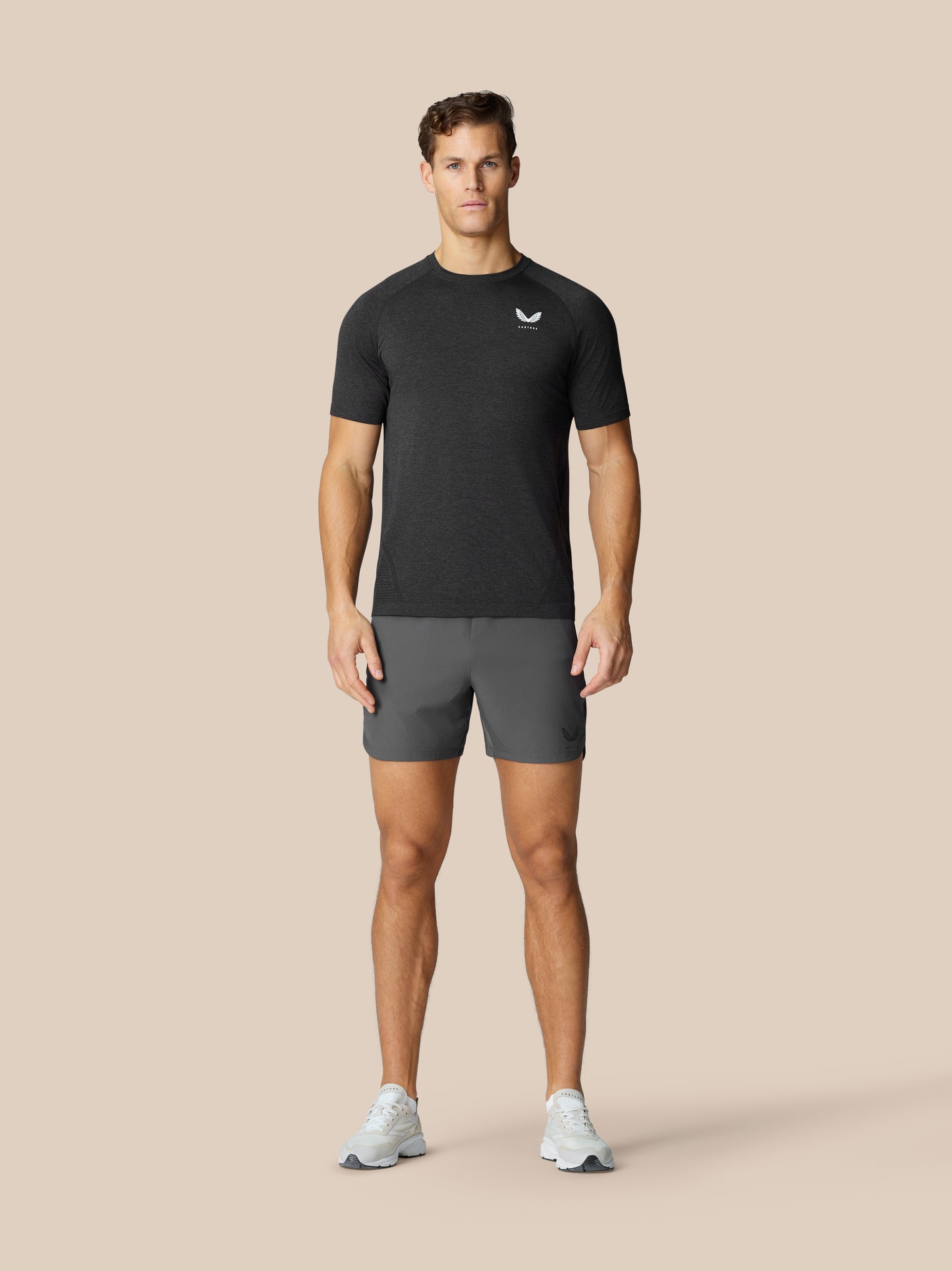 Male model wearing Flow Training Shorts in Gunmetal with a dark top, standing against a beige studio backdrop.