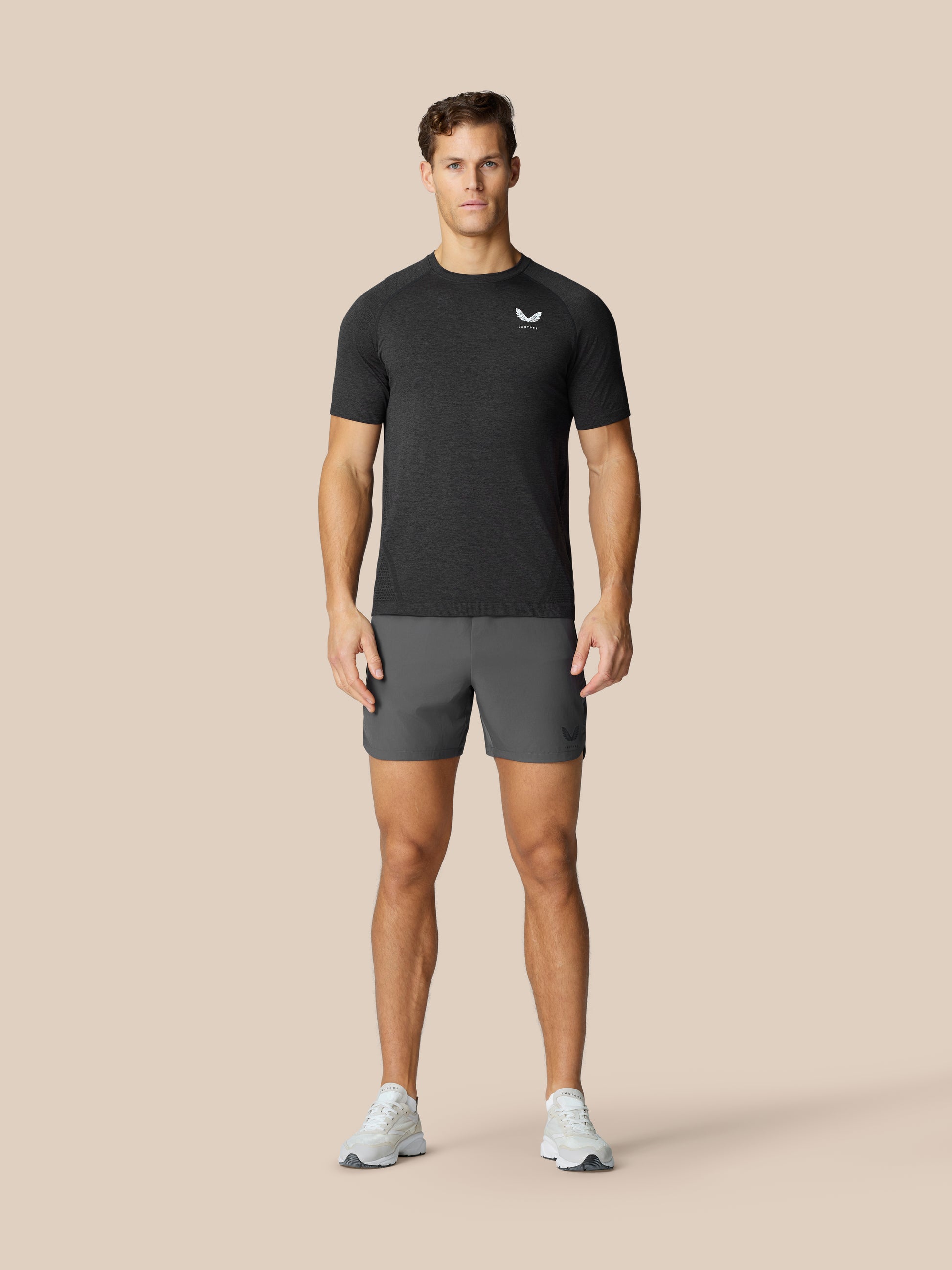 Male model wearing Flow Training Shorts in Gunmetal with a dark top, standing against a beige studio backdrop.